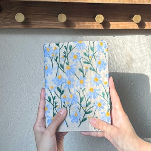 May include: A beige linen notebook cover with blue and yellow embroidered flowers. The cover is held in two hands.