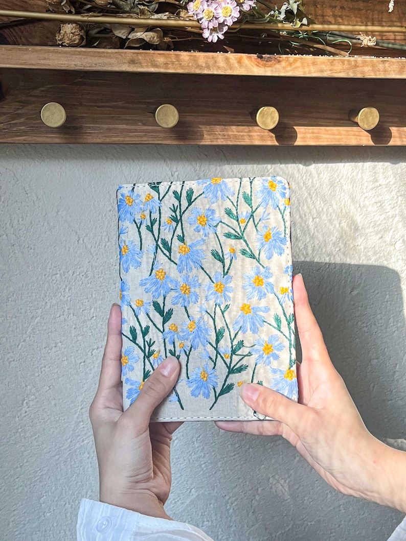 May include: A beige linen notebook cover with blue and yellow embroidered flowers. The cover is held in two hands.