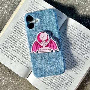 May include: A denim blue phone case with a sticker featuring a pink crescent moon, mountains, and bat wings. The sticker has the text "To the stars who listen and the dreams that one answered."