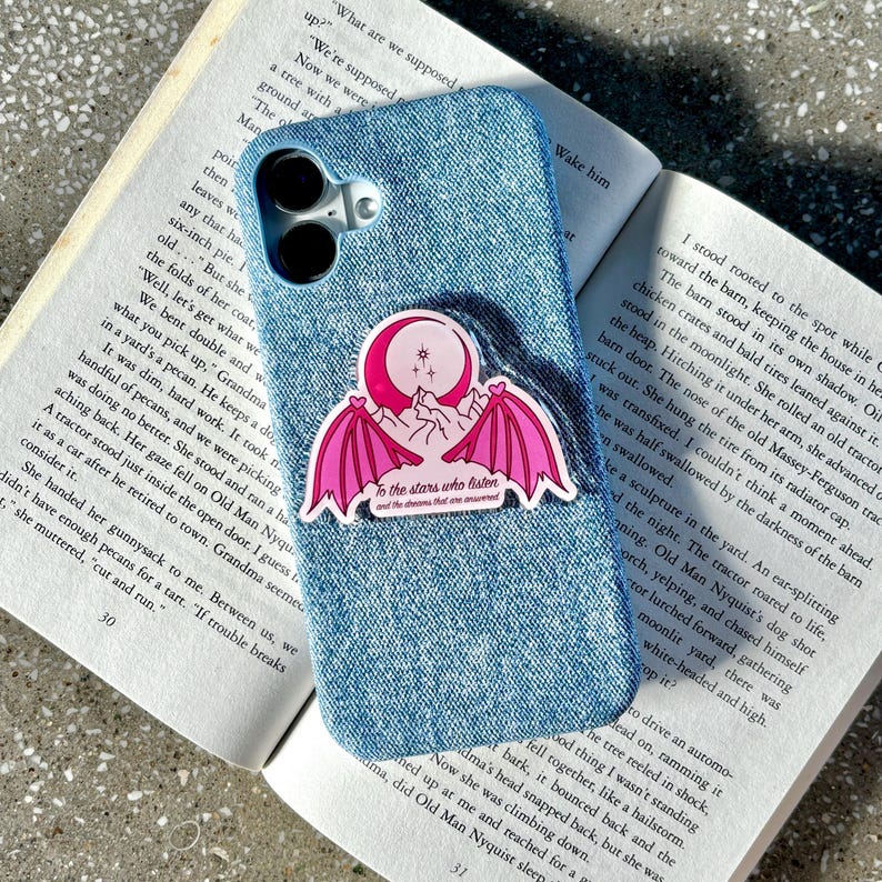 May include: A denim blue phone case with a sticker featuring a pink crescent moon, mountains, and bat wings. The sticker has the text "To the stars who listen and the dreams that one answered."