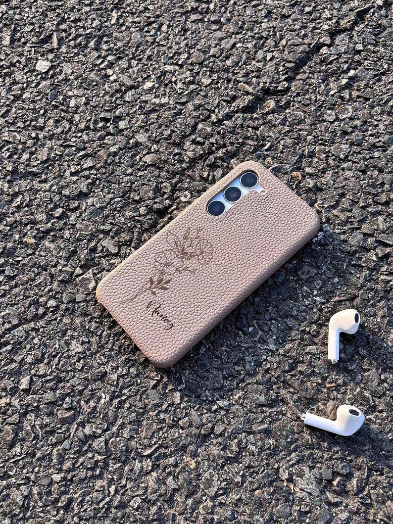 Puede incluir: Una funda de tel&eacute;fono de color marr&oacute;n claro con un dise&ntilde;o floral y la palabra "bloom" grabada en ella. La funda est&aacute; sobre una superficie de asfalto gris con dos auriculares inal&aacute;mbricos blancos cerca.