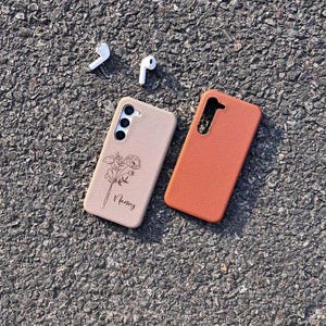 Puede incluir: Dos fundas para tel&eacute;fono, una beige con un dise&ntilde;o floral y la palabra "Nancy" grabada, y otra naranja, sobre una superficie de asfalto gris. Tambi&eacute;n se ven dos auriculares inal&aacute;mbricos blancos.