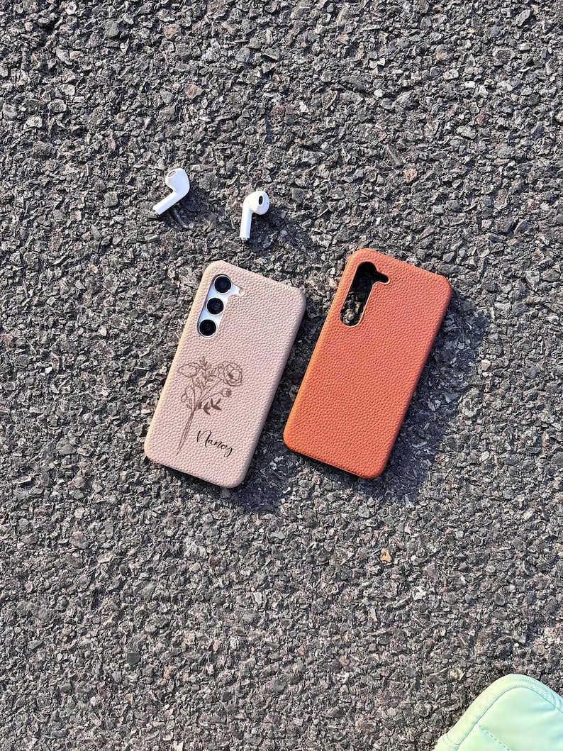 Puede incluir: Dos fundas para tel&eacute;fono, una beige con un dise&ntilde;o floral y la palabra "Nancy" grabada, y otra naranja, sobre una superficie de asfalto gris. Tambi&eacute;n se ven dos auriculares inal&aacute;mbricos blancos.