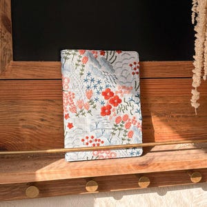 以下が含まれることがあります： 赤い、ピンク、青い花が刺繍された白いリネン製のノートカバー。カバーは金色のフックが付いた木製の棚の上に置かれています。