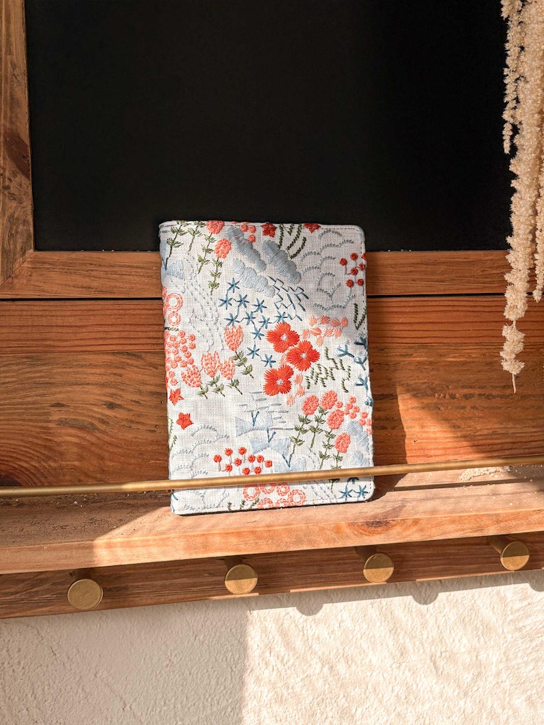 以下が含まれることがあります： 赤い、ピンク、青い花が刺繍された白いリネン製のノートカバー。カバーは金色のフックが付いた木製の棚の上に置かれています。