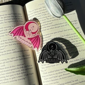 May include: Two stickers with a bat wing design, a crescent moon, and the text "To the stars who listen and the dreams that are answered." One is pink, the other black, both on an open book.