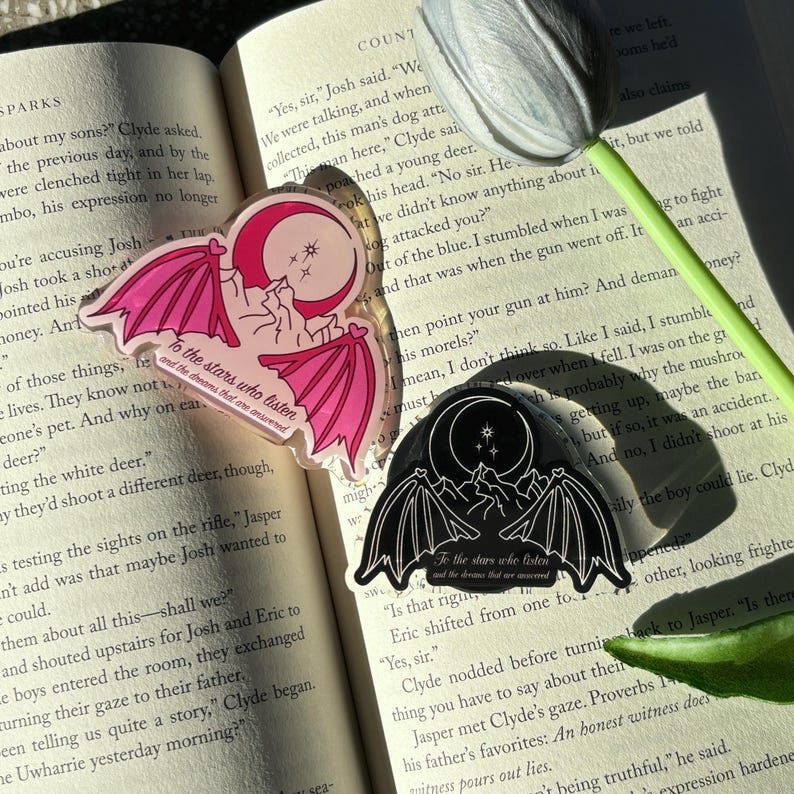 May include: Two stickers with a bat wing design, a crescent moon, and the text "To the stars who listen and the dreams that are answered." One is pink, the other black, both on an open book.