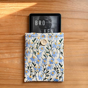 May include: A beige fabric e-reader case with a blue floral pattern and a button closure. The case is holding a black e-reader with the word "BROKEN" displayed on the screen.