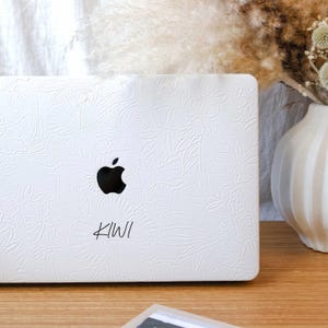 May include: White laptop with a floral design and the word "KIWI" written on it. The laptop has an Apple logo.