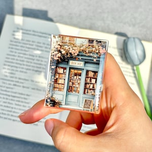 May include: A rectangular phone grip featuring a miniature illustration of a quaint blue bookshop with floral accents and the text "BOOK SHOP". The image is held by a hand, with a book in the background.