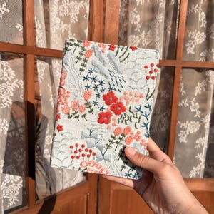 以下が含まれることがあります： 赤、ピンク、青の花が刺繍された、白いリネンのパスポートケースです。