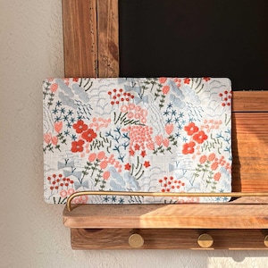 以下が含まれることがあります： 赤、ピンク、青の花が刺繍された白い布製のクラッチバッグ。クラッチバッグは真鍮製のフックが付いた木製の棚の上に置かれています。