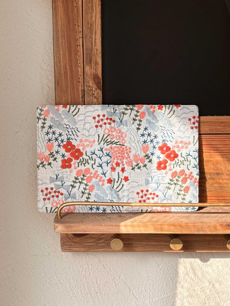 以下が含まれることがあります： 赤、ピンク、青の花が刺繍された白い布製のクラッチバッグ。クラッチバッグは真鍮製のフックが付いた木製の棚の上に置かれています。