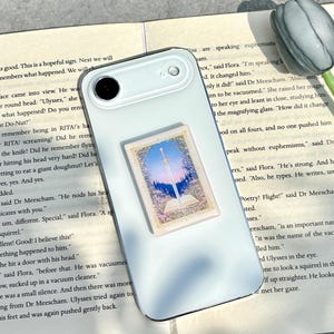 May include: A light-colored smartphone with a decorative sticker featuring a landscape scene with a sword, sky, and flowers. The phone rests on an open book with text.