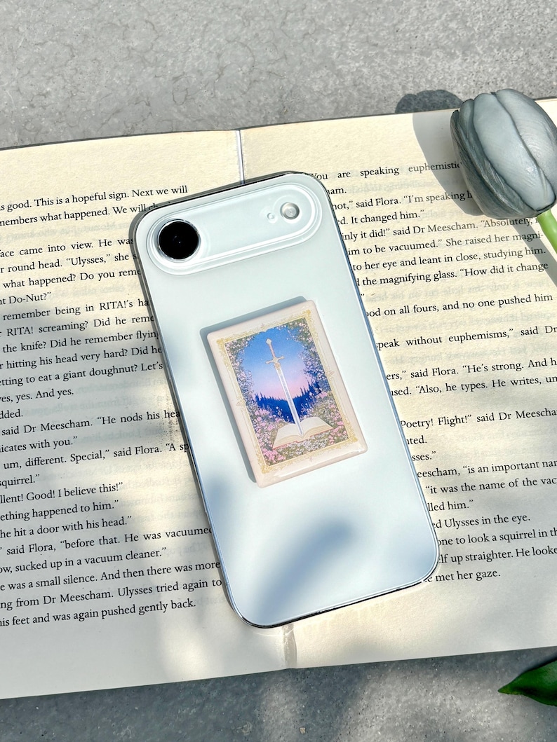 May include: A light-colored smartphone with a decorative sticker featuring a landscape scene with a sword, sky, and flowers. The phone rests on an open book with text.