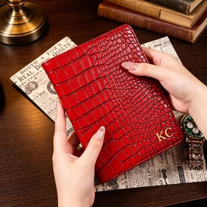 May include: A red, textured leather journal with a crocodile pattern. The journal has gold initials "KC" on the front. The journal is held over a vintage newspaper. A gold-toned watch with a brown leather band is visible.