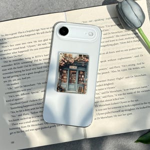 May include: White smartphone case with a rectangular sticker featuring a bookstore facade with flowers. The sticker has the text "BOOK SHOP". The phone rests on a book with text.