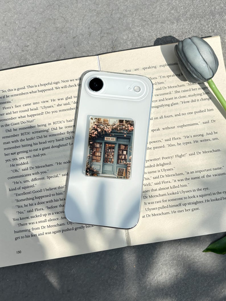 May include: White smartphone case with a rectangular sticker featuring a bookstore facade with flowers. The sticker has the text "BOOK SHOP". The phone rests on a book with text.