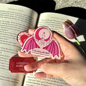 May include: A clear acrylic sticker featuring a pink bat-wing design with a crescent moon and mountain scene. The text "To the stars who listen and the dreams that are answered" is printed below the design. A red heart-shaped sticker is also visible.