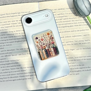 May include: A white phone case with a floral design. The case features a rectangular pop-up grip with a colorful illustration of flowers and books. The phone case is resting on an open book.