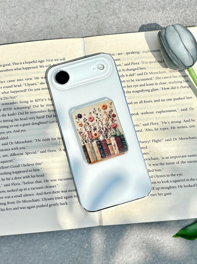 May include: A white phone case with a floral design. The case features a rectangular pop-up grip with a colorful illustration of flowers and books. The phone case is resting on an open book.