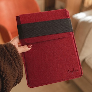 May include: A deep red felt tablet sleeve featuring a wide black elastic band closure across the top flap. The bottom right corner of the sleeve is personalized with the name Dylan embossed in a dark, cursive script font on the felt material.