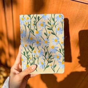 May include: A beige linen notebook cover with blue and yellow embroidered flowers. The cover has a subtle texture and a stitched edge.