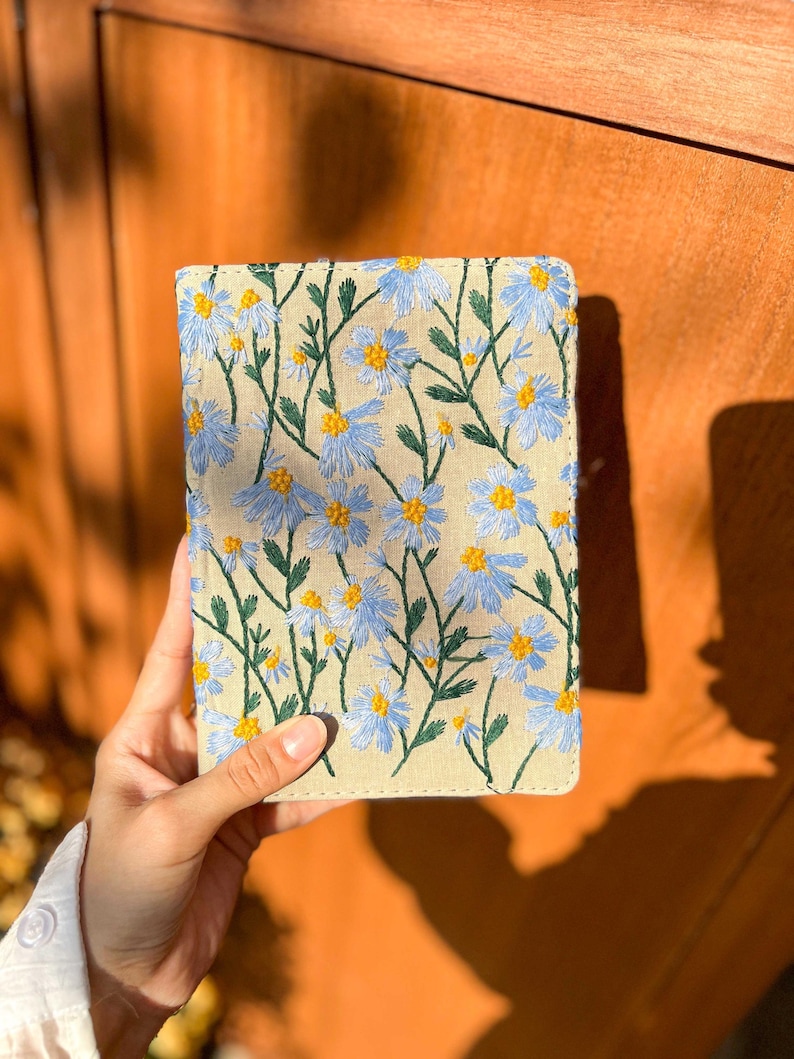 May include: A beige linen notebook cover with blue and yellow embroidered flowers. The cover has a subtle texture and a stitched edge.