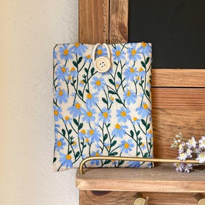 May include: A white fabric phone case with a blue floral pattern and a white button closure.