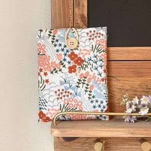 May include: A white fabric phone case with a floral embroidery design in shades of pink, orange, blue, and green. The case has a wooden button closure.