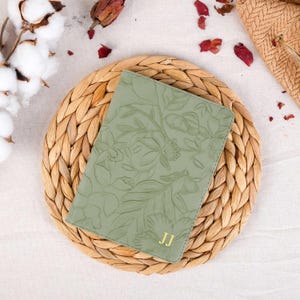 May include: A sage green leather journal with a floral embossed design and the initials "JJ" in gold lettering. The journal is sitting on a woven straw mat.