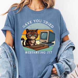 May include: Blue jean t-shirt with a graphic of a grumpy cat sitting in front of a computer. The text on the shirt reads "HAVE YOU TRIED RESTARTING IT?" The cat is brown and black with a stern expression.