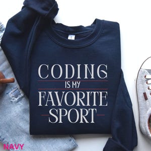 May include: Navy blue sweatshirt with the text "CODING IS MY FAVORITE SPORT" in white. The sweatshirt is folded on top of a pair of blue jeans. A brown mug and a gray sneaker are visible in the background.