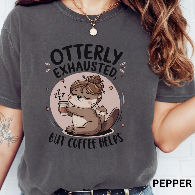 May include: A gray t-shirt with the text "Otterly Exhausted, But Coffee Helps" and a cartoon illustration of an otter holding a cup of coffee. The otter is wearing a pink headband and has a small otter on its back.