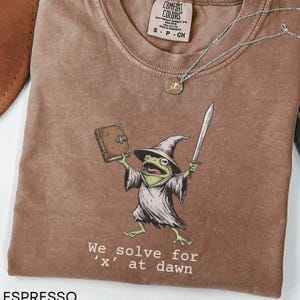 May include: Espresso-colored t-shirt with a whimsical graphic of a frog wizard holding a book and sword. The text "We solve for 'x' at dawn" is printed below the frog. A silver necklace with a pendant is also visible.