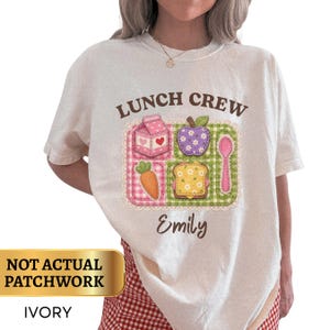 May include: An ivory short-sleeve t-shirt featuring the text LUNCH CREW and the name Emily in brown. The graphic shows a divided lunch tray with a pink milk carton, a purple apple, a carrot, a sandwich, and a pink spoon in a patchwork style.