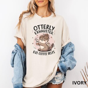 May include: A white t-shirt with the text "Otterly Exhausted, But Coffee Helps" and a cartoon otter holding a cup of coffee. The otter has a baby otter on its back. The shirt is available in ivory.