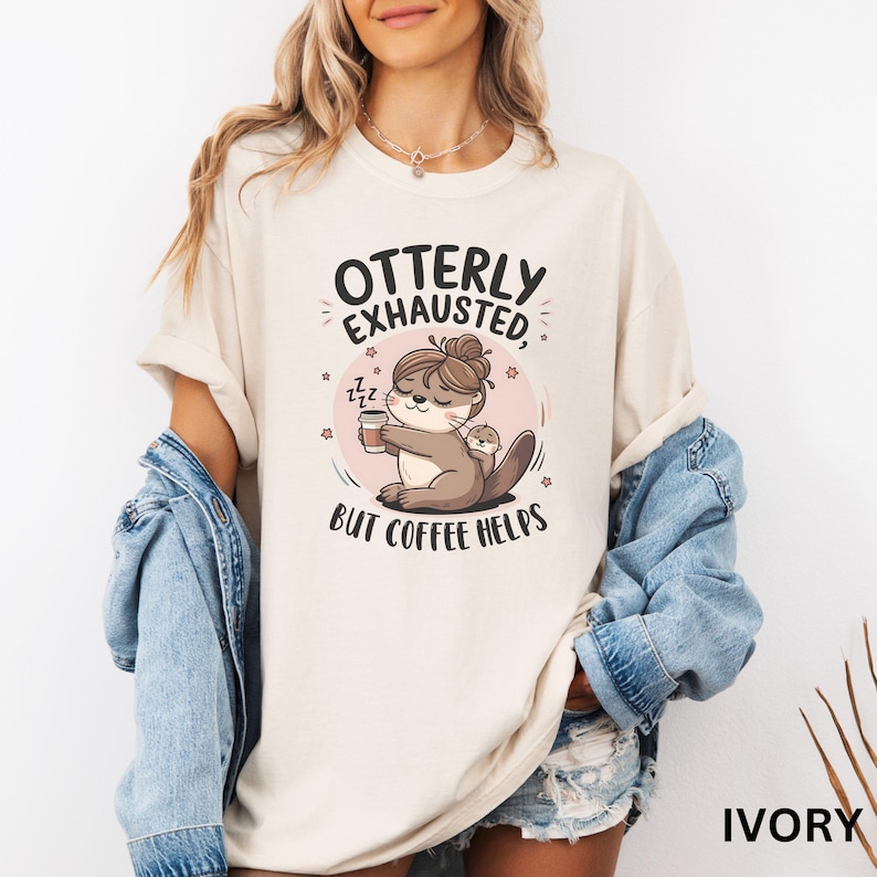 May include: A white t-shirt with the text "Otterly Exhausted, But Coffee Helps" and a cartoon otter holding a cup of coffee. The otter has a baby otter on its back. The shirt is available in ivory.