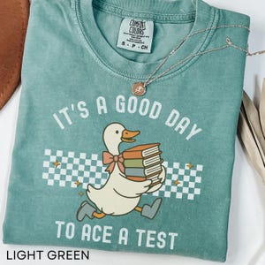May include: Light green t-shirt with the text "IT'S A GOOD DAY TO ACE A TEST". The graphic features a cartoon duck carrying books. The shirt has a crew neck and short sleeves. A gold necklace with a pendant is draped across the shirt.