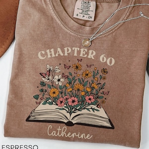 May include: Espresso-colored t-shirt with the words "CHAPTER 60" and "Catherine" printed on it. The shirt features a floral design with butterflies and an open book. A silver necklace with a pendant is also visible.