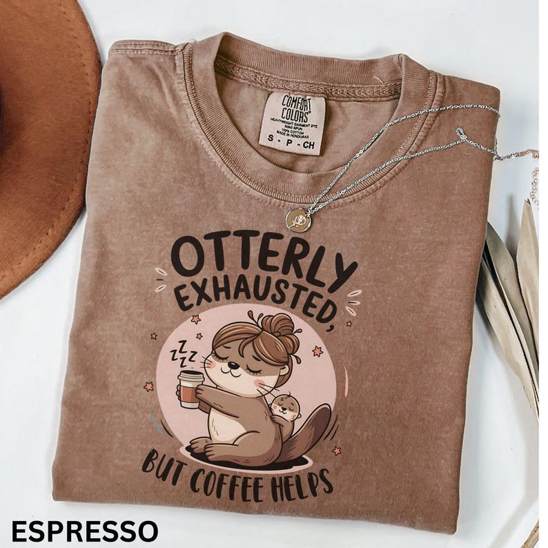 May include: A brown t-shirt with a graphic of an otter holding a cup of coffee and the text "OTTERLY EXHAUSTED, BUT COFFEE HELPS".
