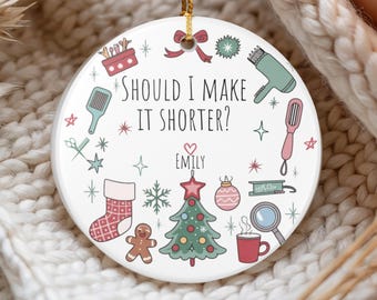 May include: A white ceramic Christmas ornament with a gold string for hanging. The ornament features a festive design with a Christmas tree, stocking, and the text "SHOULD I MAKE IT SHORTER?" with the name "EMILY".