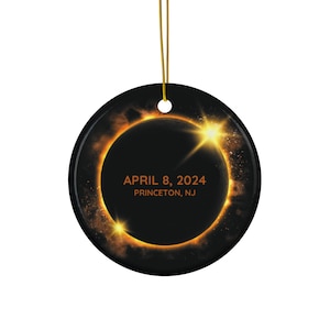 May include: A round black ceramic ornament featuring a graphic of a solar eclipse with a glowing orange ring of light. The text "APRIL 8, 2024" and "PRINCETON, NJ" is printed in orange in the center. A gold string is attached to the top for hanging.