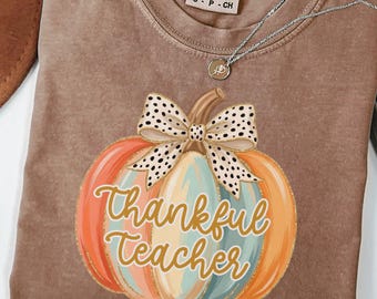 May include: An espresso-colored t-shirt featuring a graphic of a multi-colored pumpkin with a white and black polka-dot bow. The text Thankful Teacher is written in gold cursive across the pumpkin. A silver chain necklace rests on the neckline.