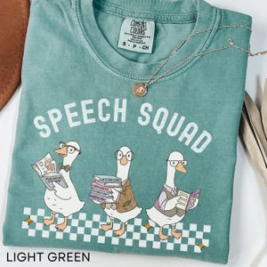 May include: Light green t-shirt with the words "SPEECH SQUAD" in white. The shirt features a graphic of three geese wearing glasses and holding books. The bottom of the shirt has a checkered pattern. A gold necklace with a pendant is draped across the shirt.