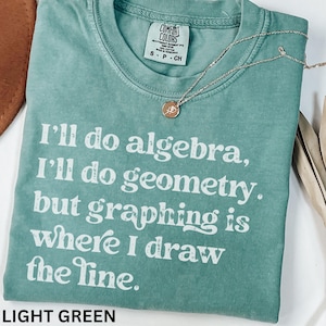 May include: Light green t-shirt with the text "I'll do algebra, I'll do geometry, but graphing is where I draw the line." A gold necklace with a pendant rests on the shirt. The shirt has a Comfort Colors tag.