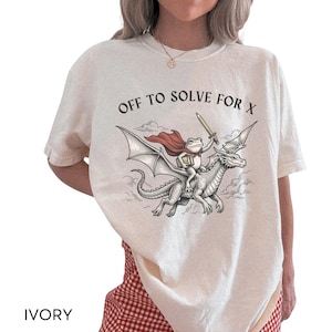 May include: An ivory t-shirt featuring a graphic illustration of a frog wearing a red cape, holding a book and a sword, while riding a flying dragon. The text "OFF TO SOLVE FOR X" is printed in black above the illustration of the frog and dragon.