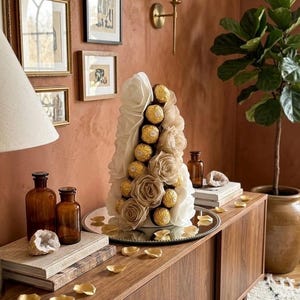 Peut inclure: Une composition décorative avec des chocolats dorés et des roses en tissu. L'arrangement est présenté sur un plateau miroir posé sur un meuble en bois. Le meuble est dans une pièce avec des œuvres d'art encadrées et une plante en pot.