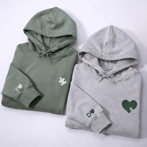 May include: Two folded hoodies, one sage green and one gray. The green hoodie has a white puzzle piece and the letter "F" with a heart on the sleeve. The gray hoodie has a green heart with a puzzle piece and the letter "D" with a heart on the sleeve.