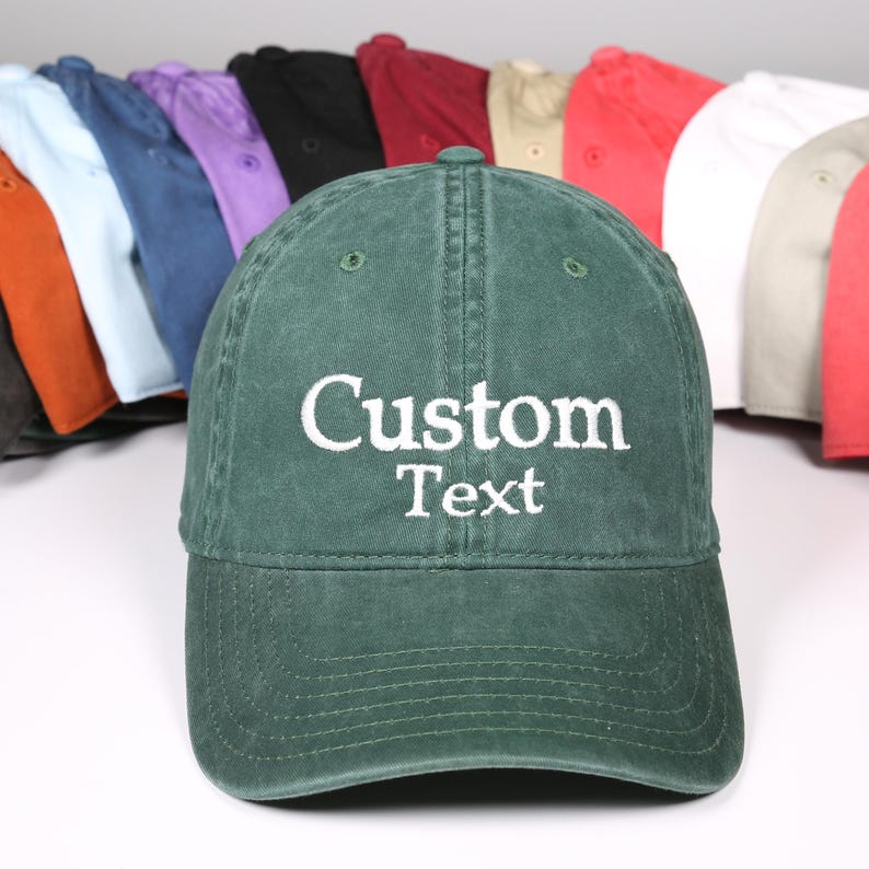 Puede incluir: Gorra de b&eacute;isbol verde con el texto "Custom Text" bordado en blanco. Se ven otras gorras de colores en el fondo. Las gorras son de varios colores, incluyendo rojo, blanco y azul.